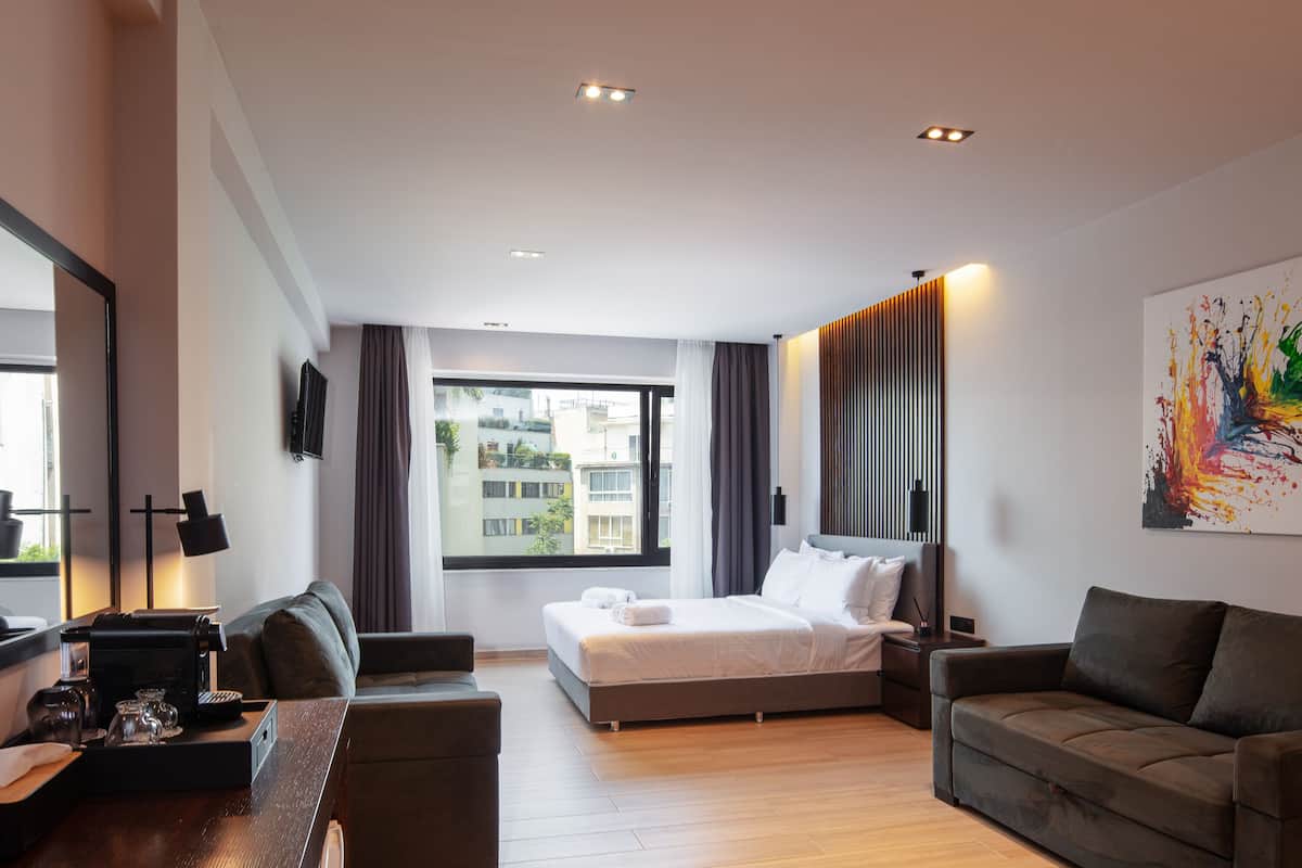 A modern hotel room with a large bed, a sofa, a desk with a lamp, and a large window with curtains.