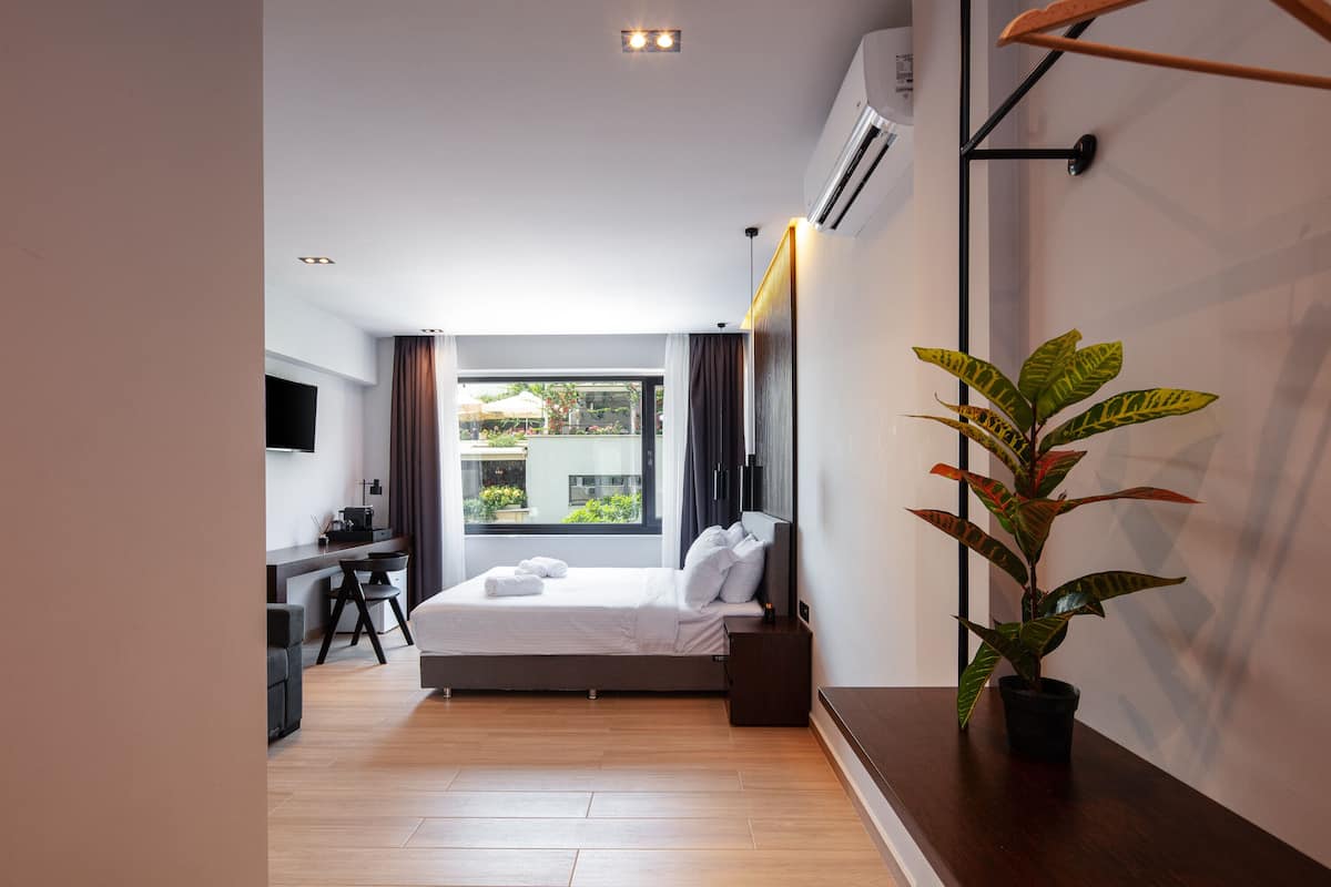 A modern hotel room with a bed, a desk, a chair, a potted plant, and a large window with curtains.