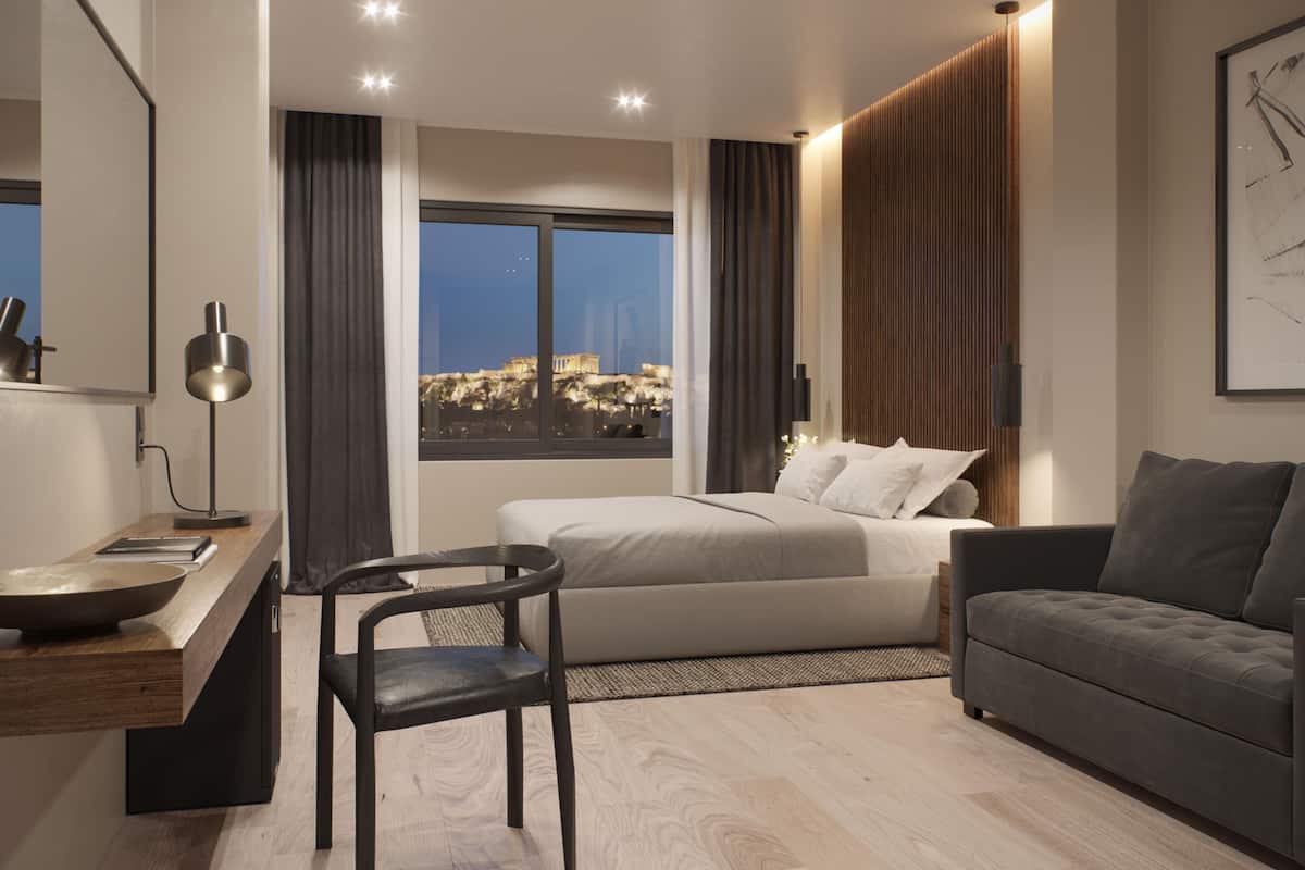 A modern hotel room with a bed, a desk, a chair, a sofa, and a large window with a view of a cityscape.