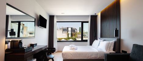 A modern hotel room with a bed, a desk, a chair, a TV, and a view of the city.