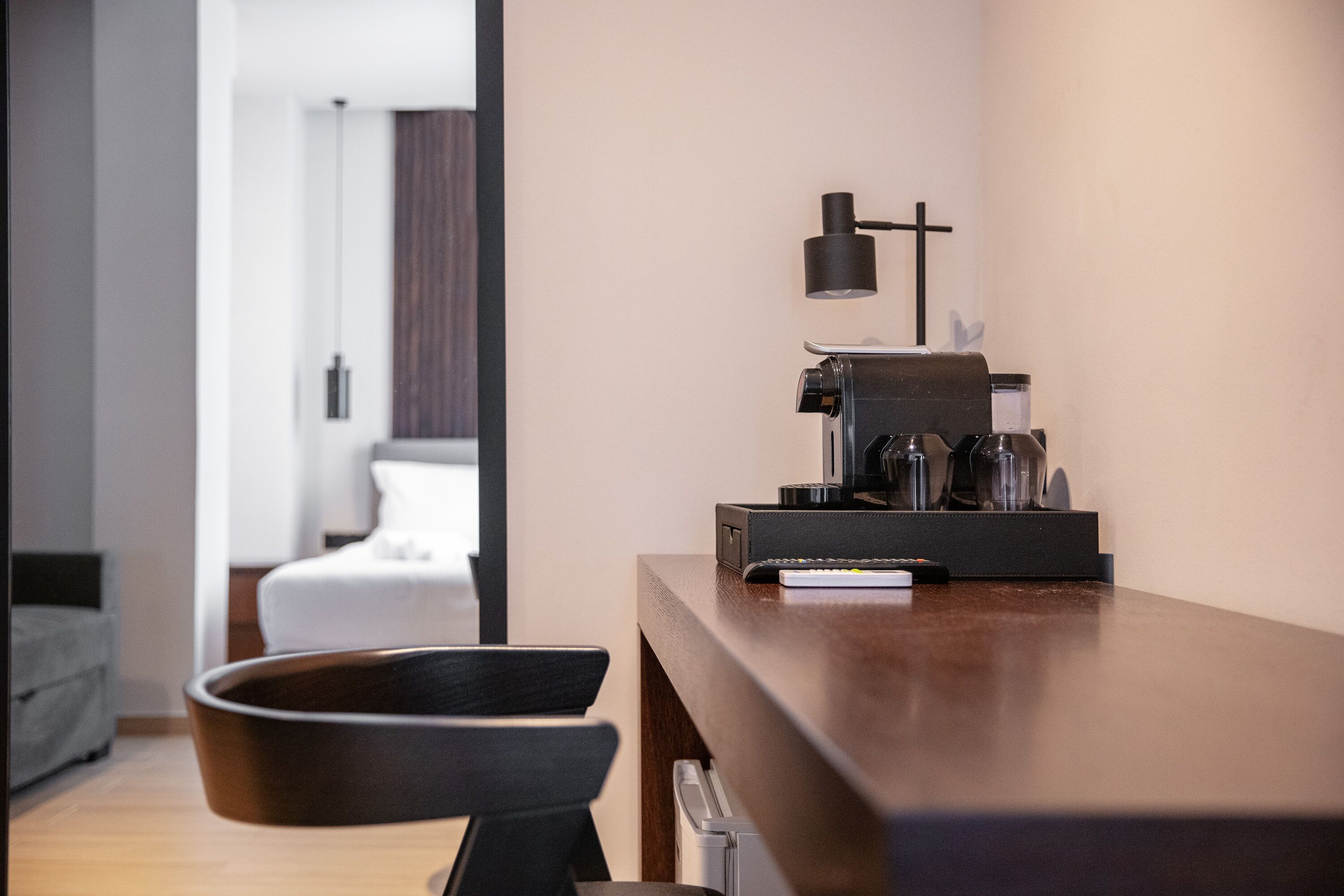 A modern hotel room with a wooden desk, a coffee machine, and a lamp. A bed and a chair are visible in the background.