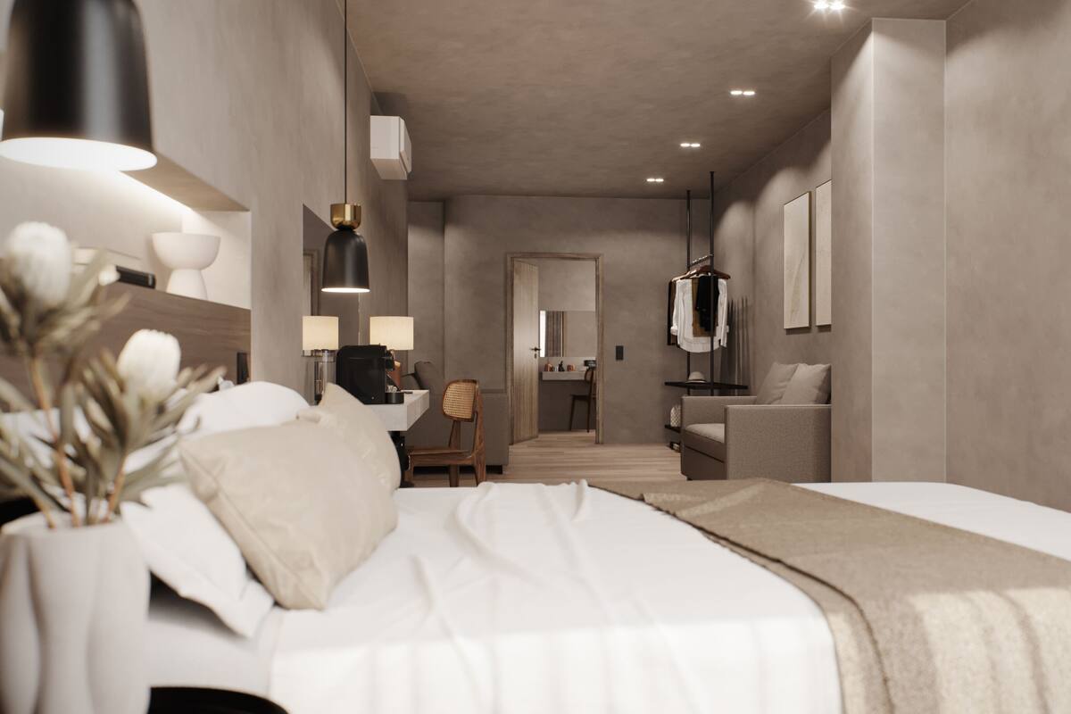 A modern hotel room with a large bed, bedside tables, a desk, and a chair. There is a mirror, a wardrobe, and a bathroom visible in the background.