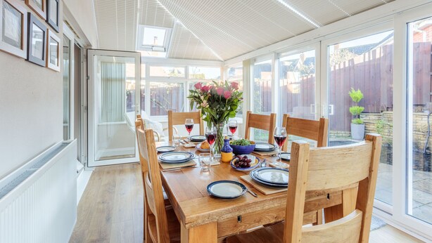 A dining area with a wooden table set for four, surrounded by chairs, with a view of the backyard through large windows.