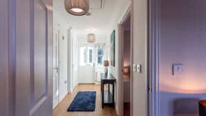 A hallway with wooden flooring, a blue rug, a table with a lamp, and a door leading to another room.