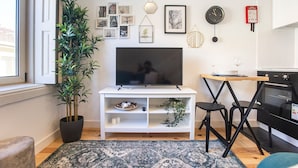 A modern living room with a TV on a white stand, a dining table with chairs, a kitchen area with an oven, and a large potted plant.