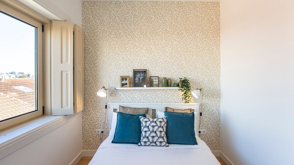 A bedroom with a bed, pillows, a shelf with decorative items, a window, and a patterned wall.