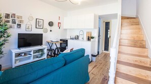 A modern living room with a teal sofa, a flat-screen TV, and a kitchen area with white cabinets and a wooden staircase.