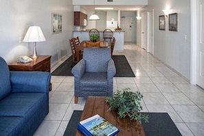 Uma sala de estar moderna com um sofá azul, uma poltrona estampada, uma mesa de centro de madeira com um livro e uma planta, e um espaço de jantar com mesa e cadeiras.