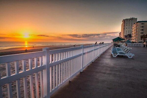Um calçadão à beira-mar com guarda-corpo branco, espreguiçadeiras e vista para o oceano ao pôr do sol.