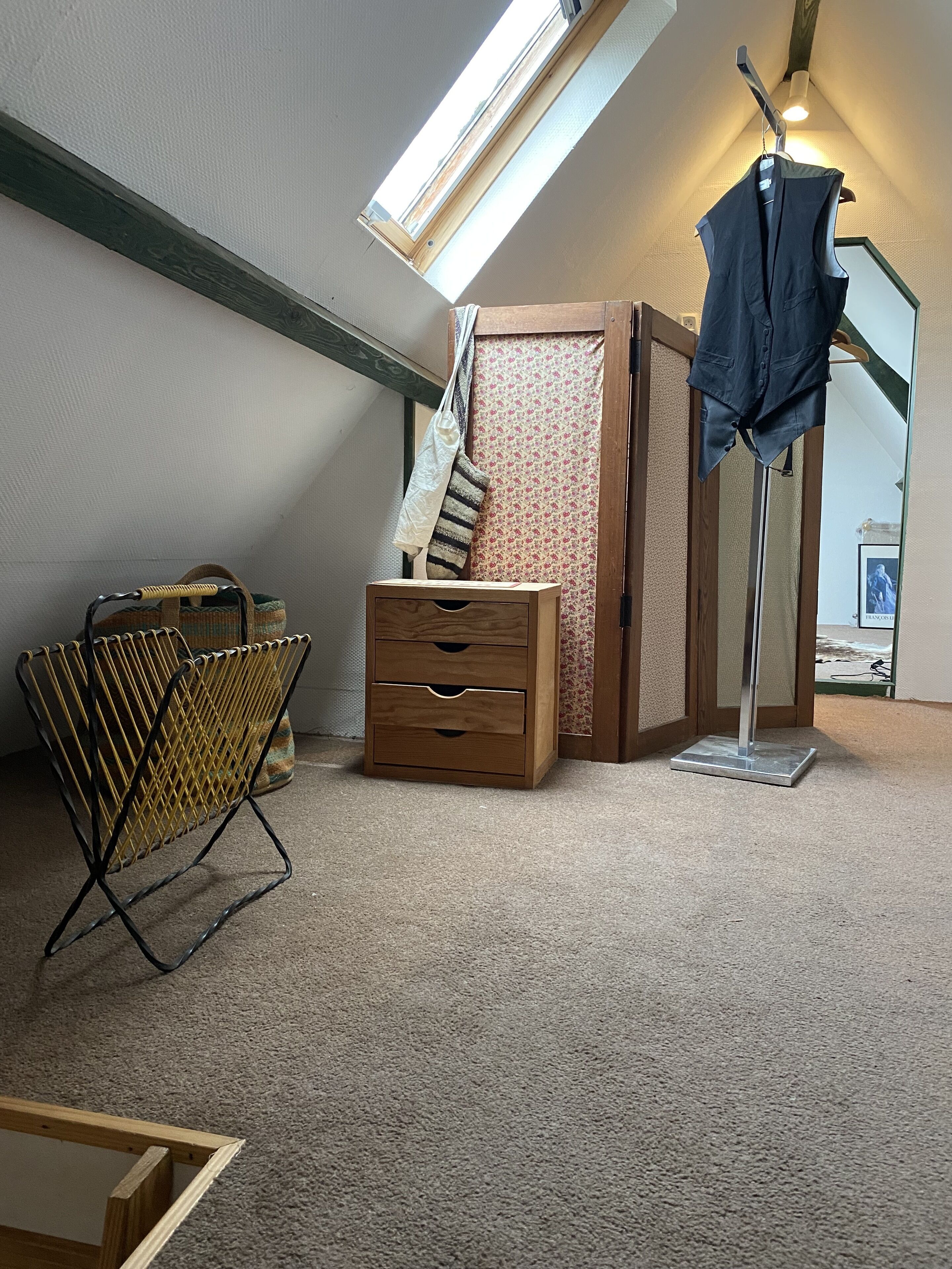 Een kamer met een schuine balkenlaag, een houten dressoir, een metalen klapstoel, een kapstok en een wenteltrap met een bloemmotief op de muur.