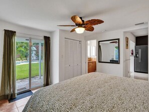 Een slaapkamer met een groot bed, een plafondventilator, een schuifdeur naar een balkon en een zichtbare keuken door een open deur.