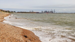 A pebbled beach with a calm sea, a distant port with cranes, and a cloudy sky.