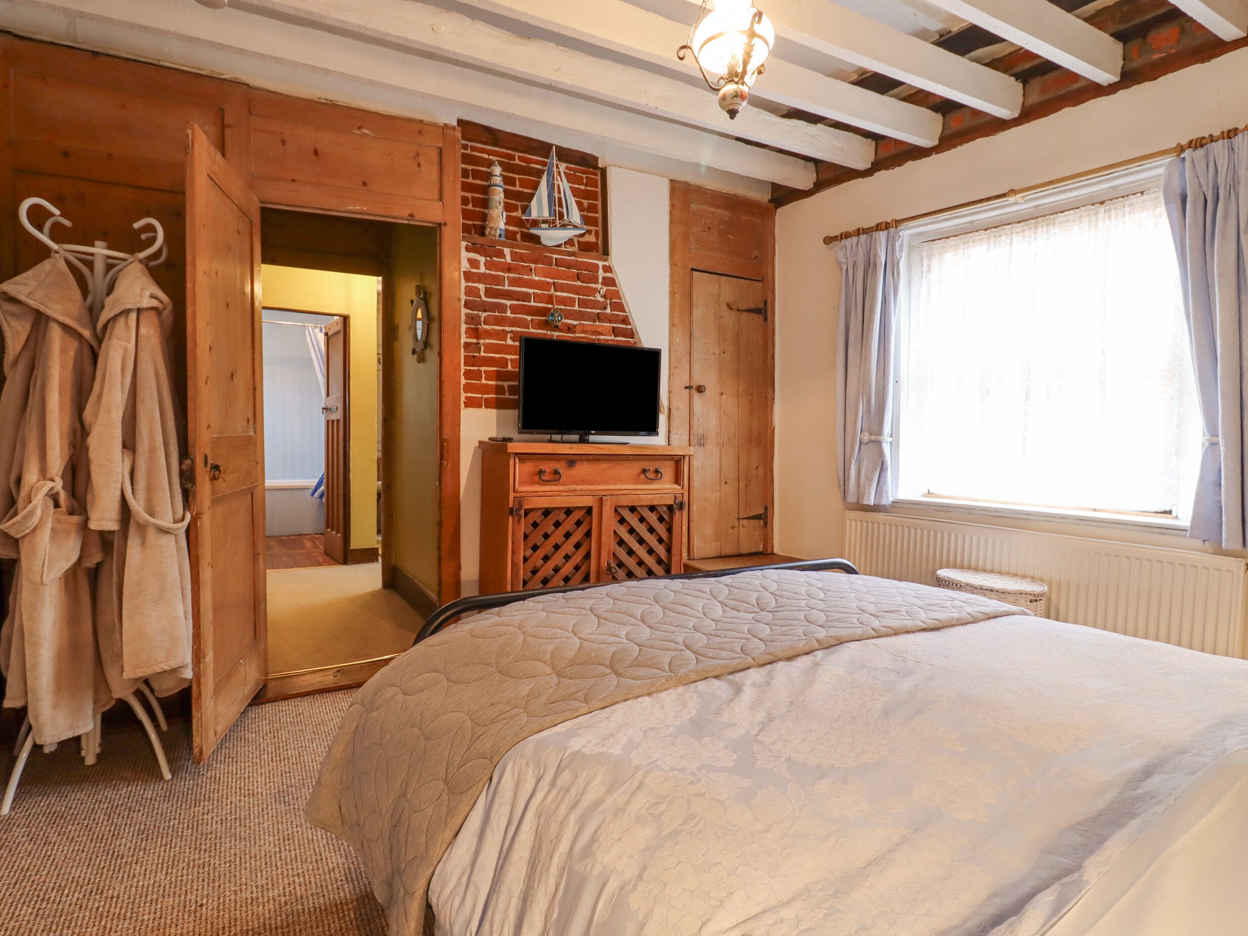 A bedroom with a bed, a TV, a wooden wardrobe, a brick wall, and a window with curtains.
