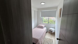 A bedroom with a bed, a small white nightstand, a window with a view of buildings, and a door.