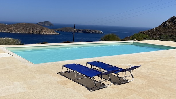 A rectangular swimming pool with clear blue water, surrounded by beige tiles. There are two blue lounge chairs with metal frames and blue cushions. The pool is set against a backdrop of rocky terrain and the sea.