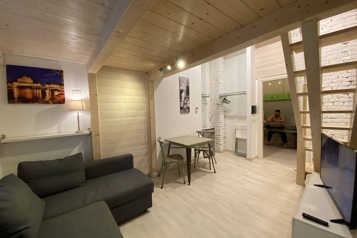 A modern living room with a grey sofa, a dining table with chairs, a TV, and a brick wall.