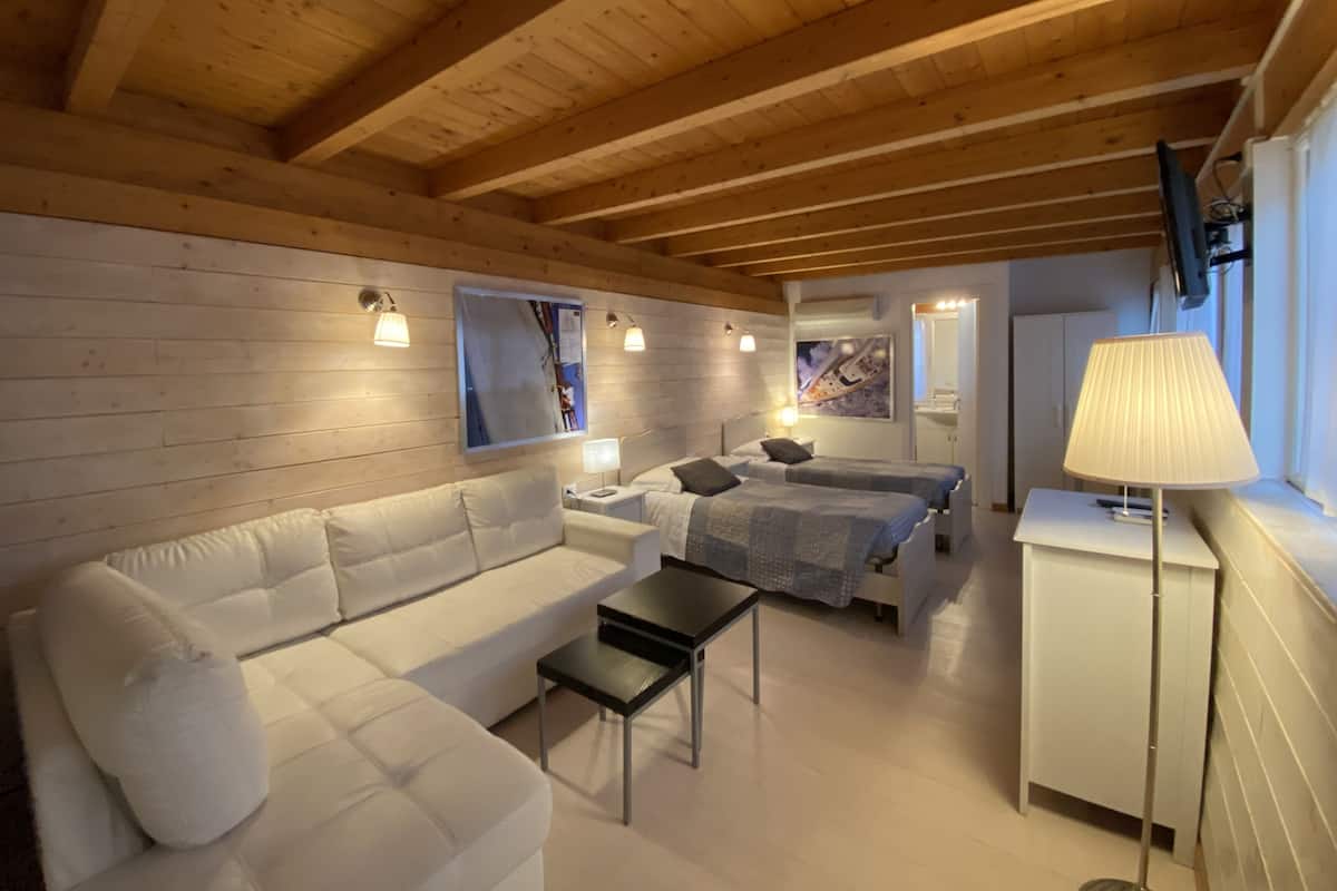 A cozy room with a wooden ceiling, a white sofa, a bed with a grey blanket, a small table, and a lamp.