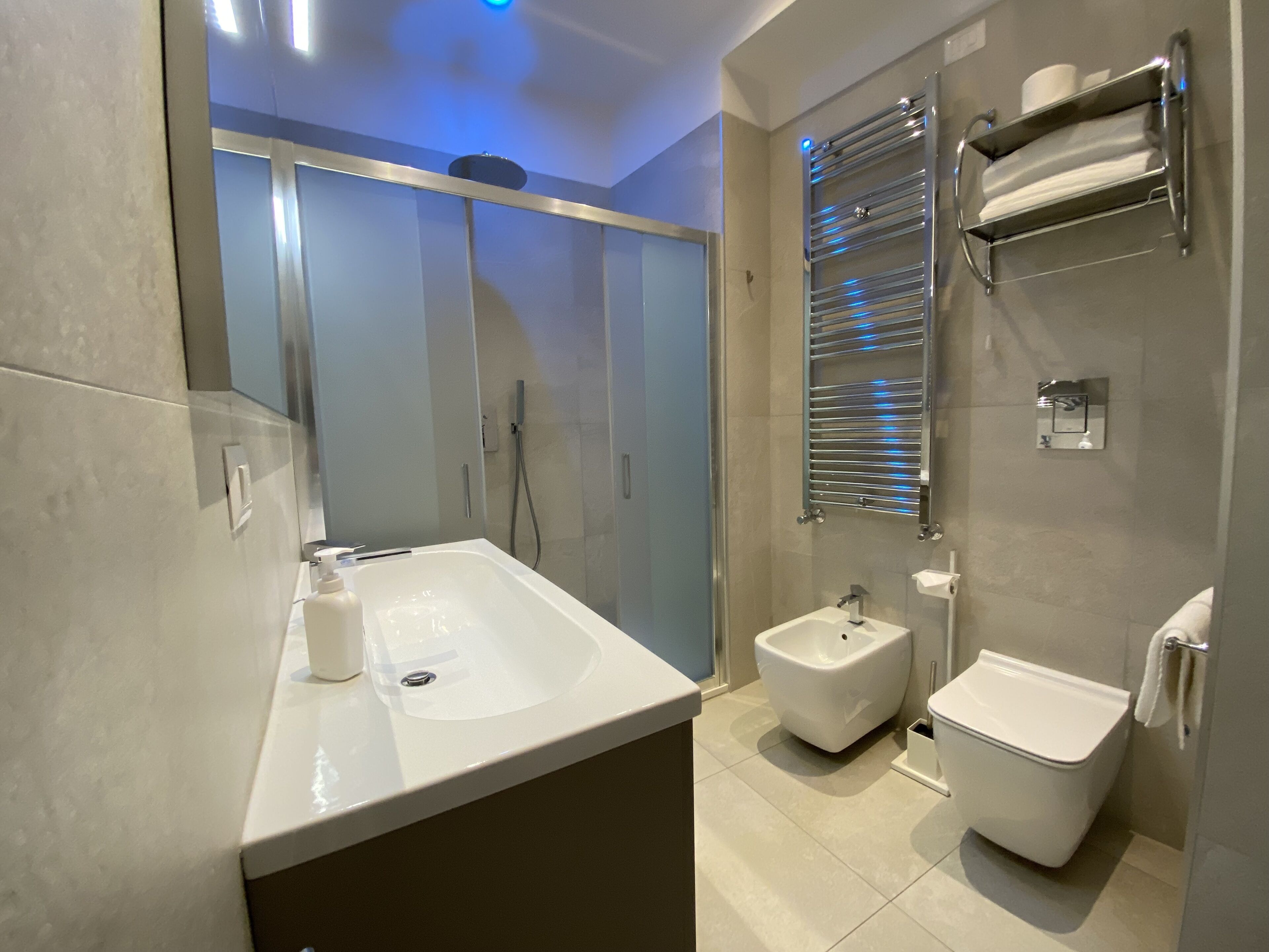 A modern bathroom with a white sink, bidet, and toilet. There is a towel rack with a mirror above the toilet.