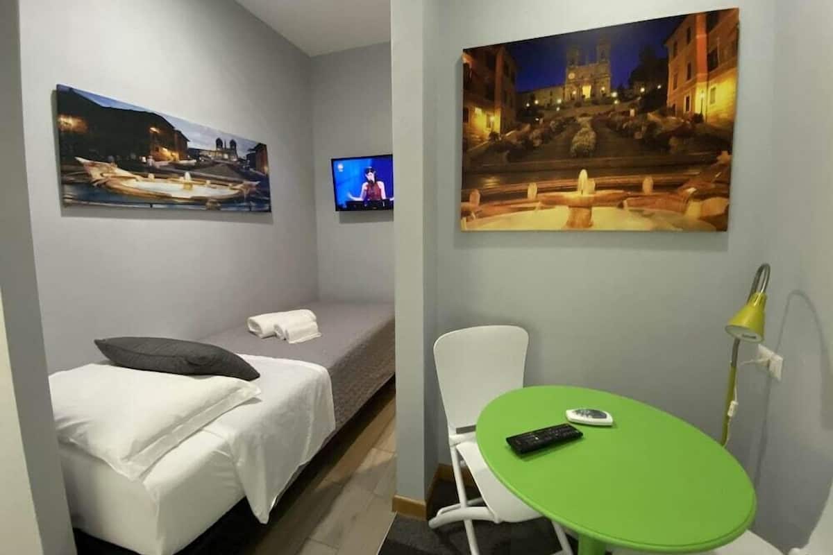 A hotel room with a bed, a green table, a TV, and two paintings on the walls.