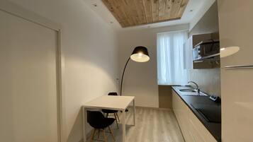 Une cuisine compacte avec un plan de travail blanc, une chaise noire, une lampe moderne et une fenêtre ornée de rideaux légers.