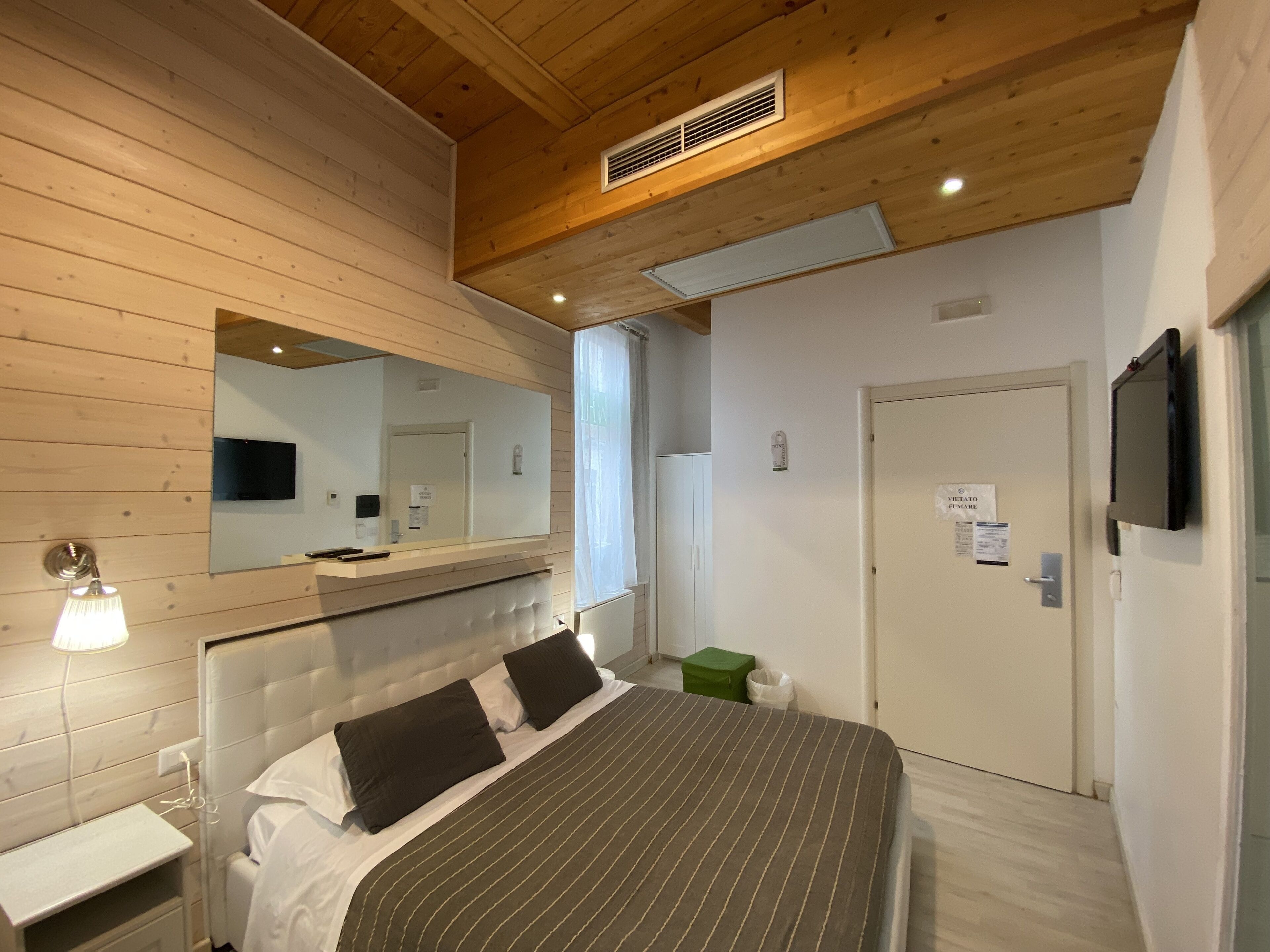 A modern bedroom with a wooden ceiling, a large bed with striped bedding, a bedside table, a mirror, and a television mounted on the wall.