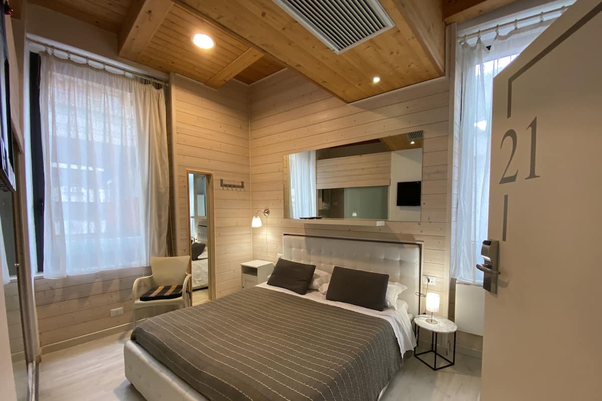 A bedroom with a large bed, a bedside table, a chair, a mirror, and a window with curtains.