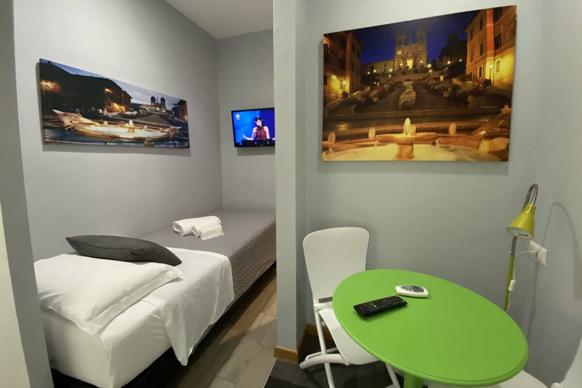 A hotel room with a bed, a green table, a chair, a TV, and two paintings on the wall.