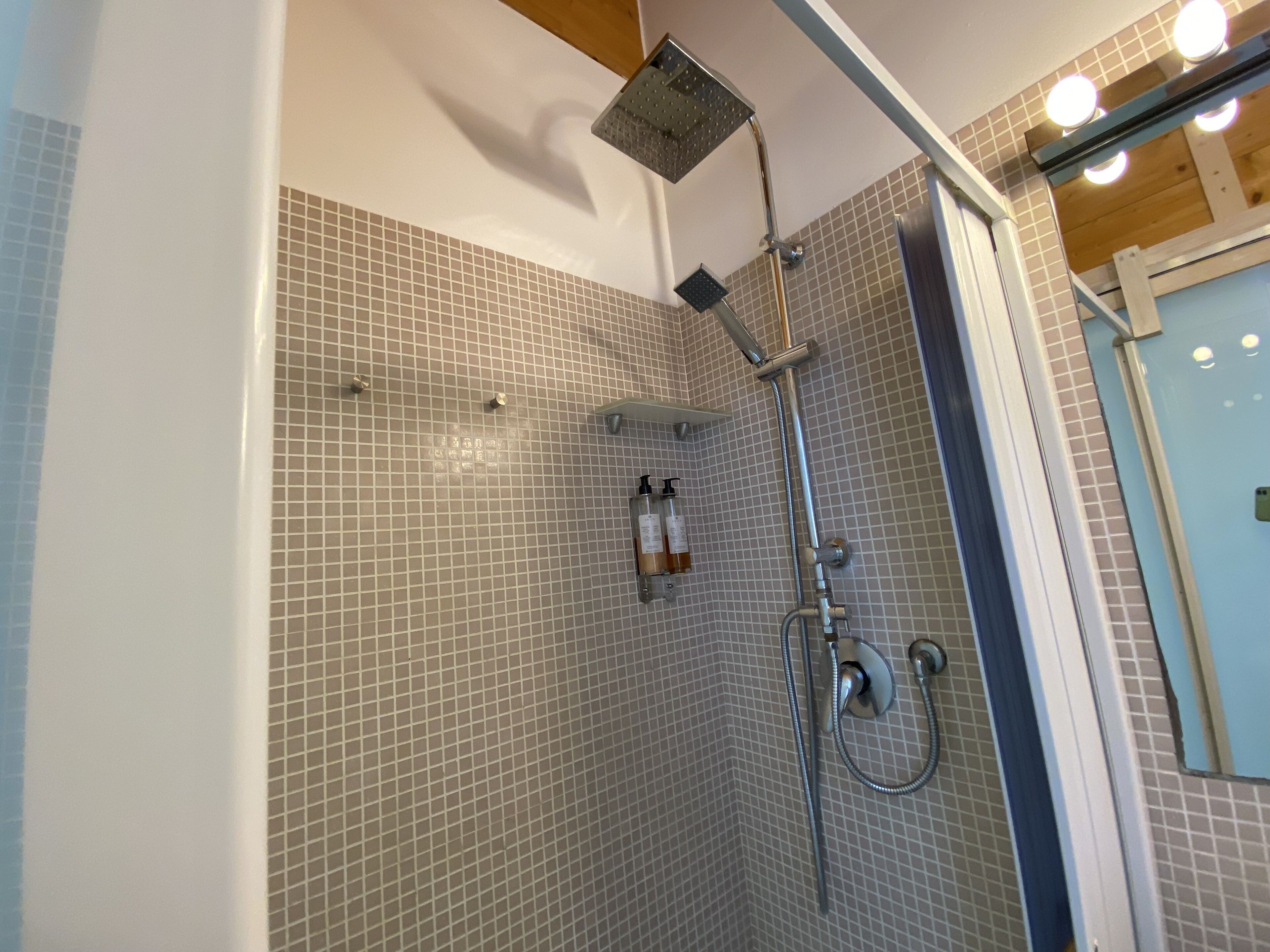A shower with a handheld showerhead and a wall-mounted showerhead, tiled walls, and a shelf with toiletries.