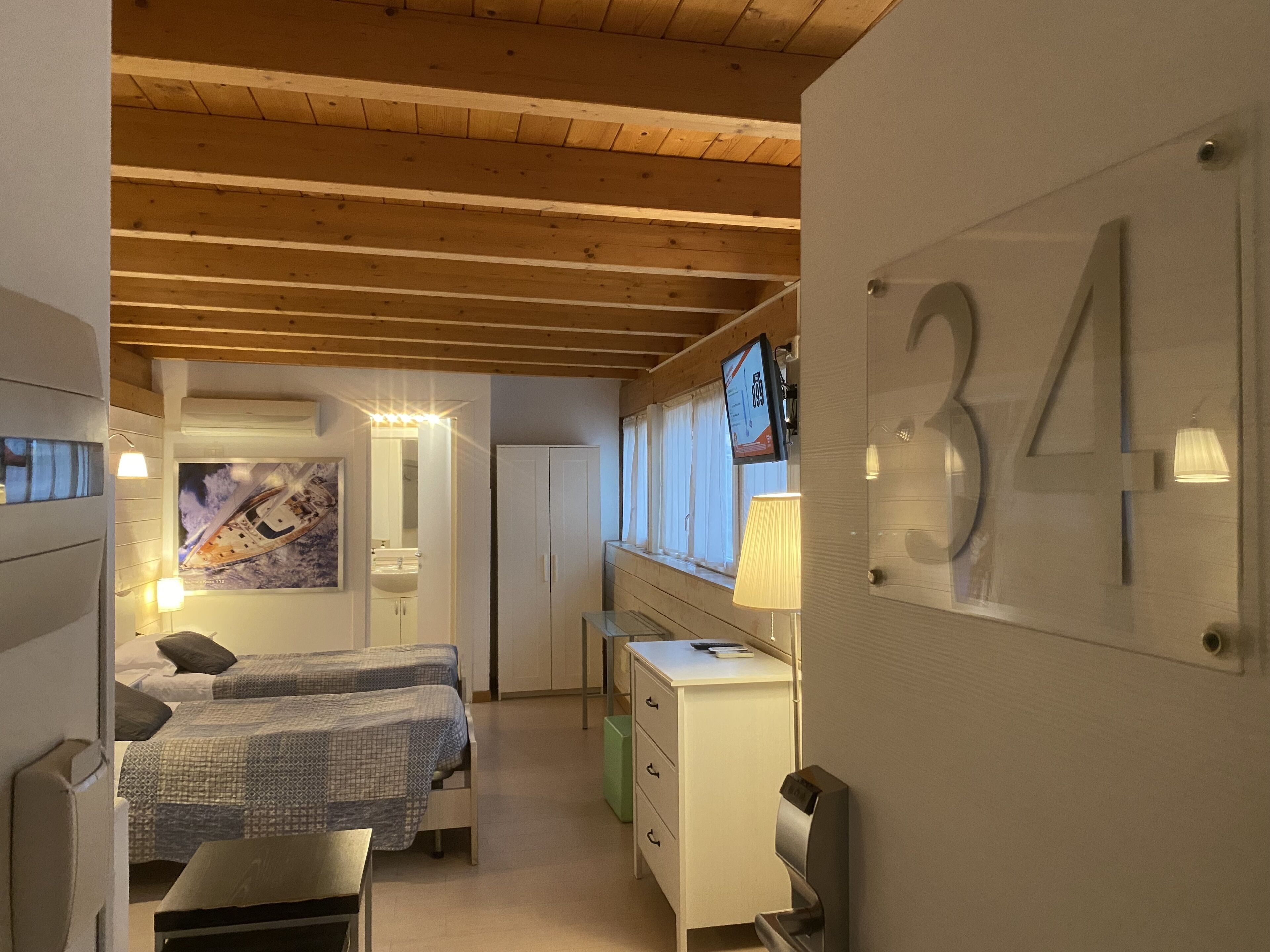 A hotel room with two beds, a desk with a computer, a TV, and a large number '34' on the wall.
