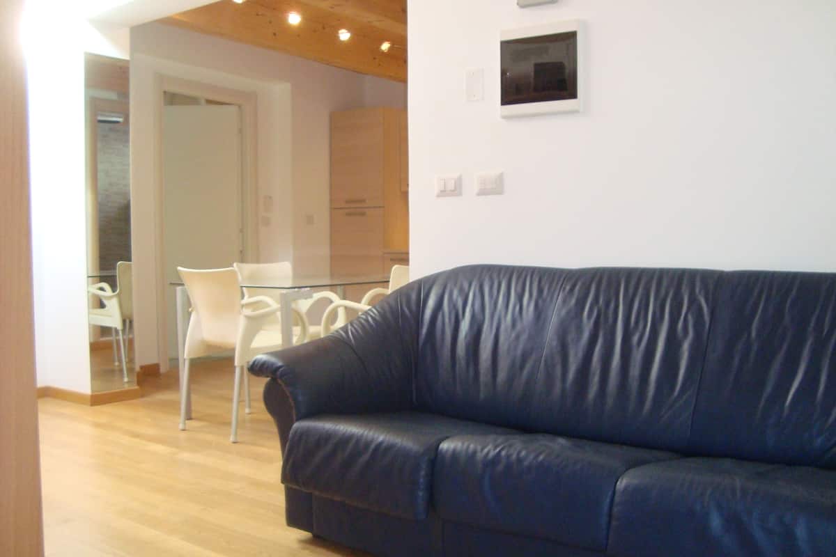 A living room with a blue leather sofa, a glass coffee table, and white chairs. There is a wooden floor and a white wall with a small picture frame.