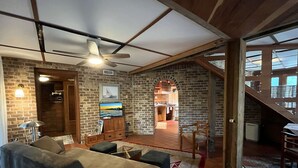 Amplia sala de estar con sofá, televisor y ventilador de techo. Se observa una pared de ladrillo y una escalera que conduce a un nivel superior.
