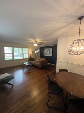 A spacious living room with hardwood flooring, a ceiling fan, a dining table with chairs, and a large window with blinds. - Stunning home in the middle of midtown close to fairgrounds. (Tulsa)