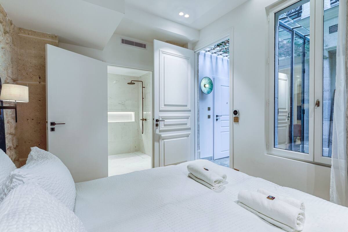 A hotel room with a large bed, a bathroom with a glass shower, and a view of the outside through a window.