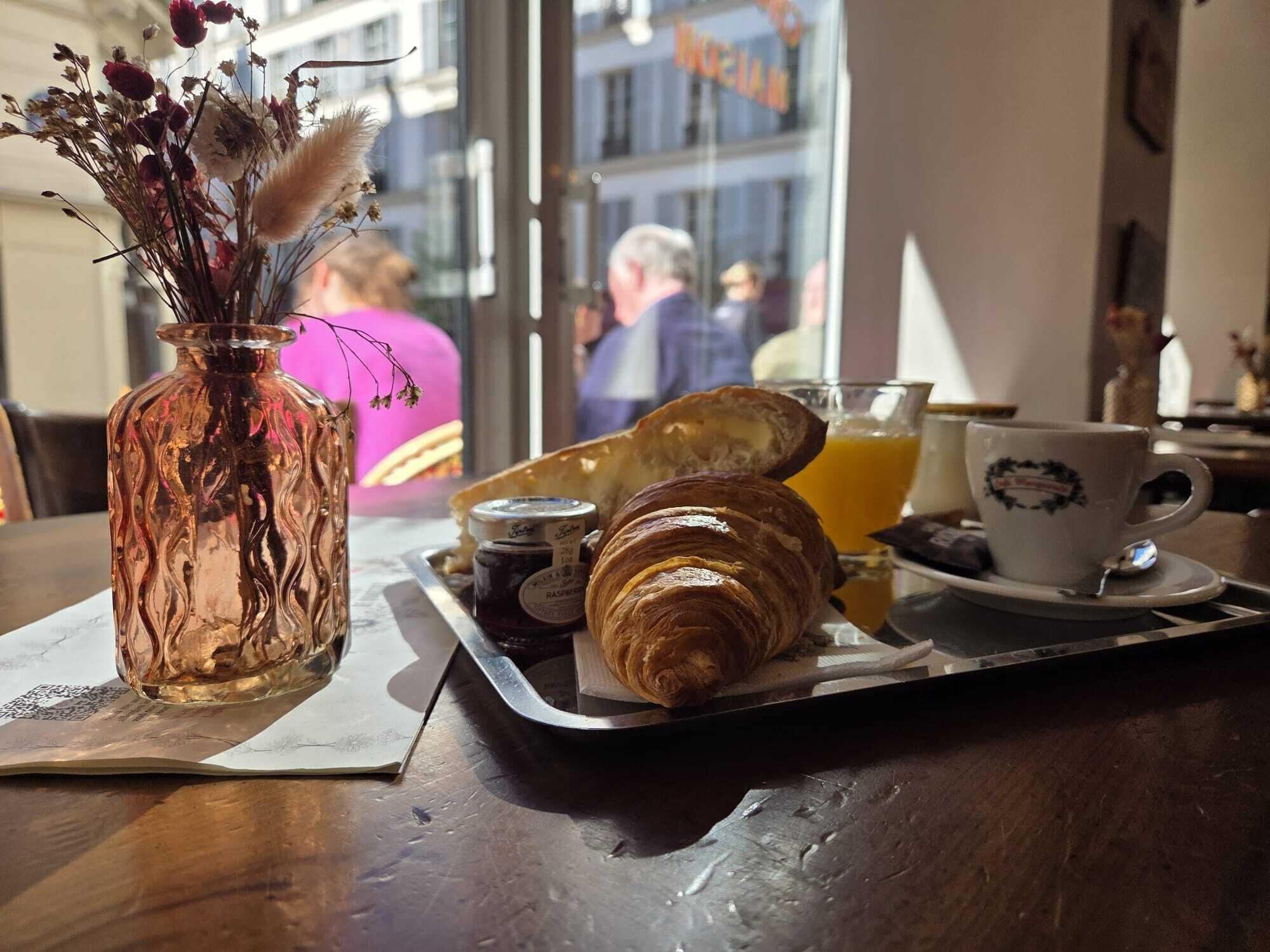 A breakfast tray with croissants, a glass of orange juice, and a cup with a logo, placed on a table with a vase of dried flowers.