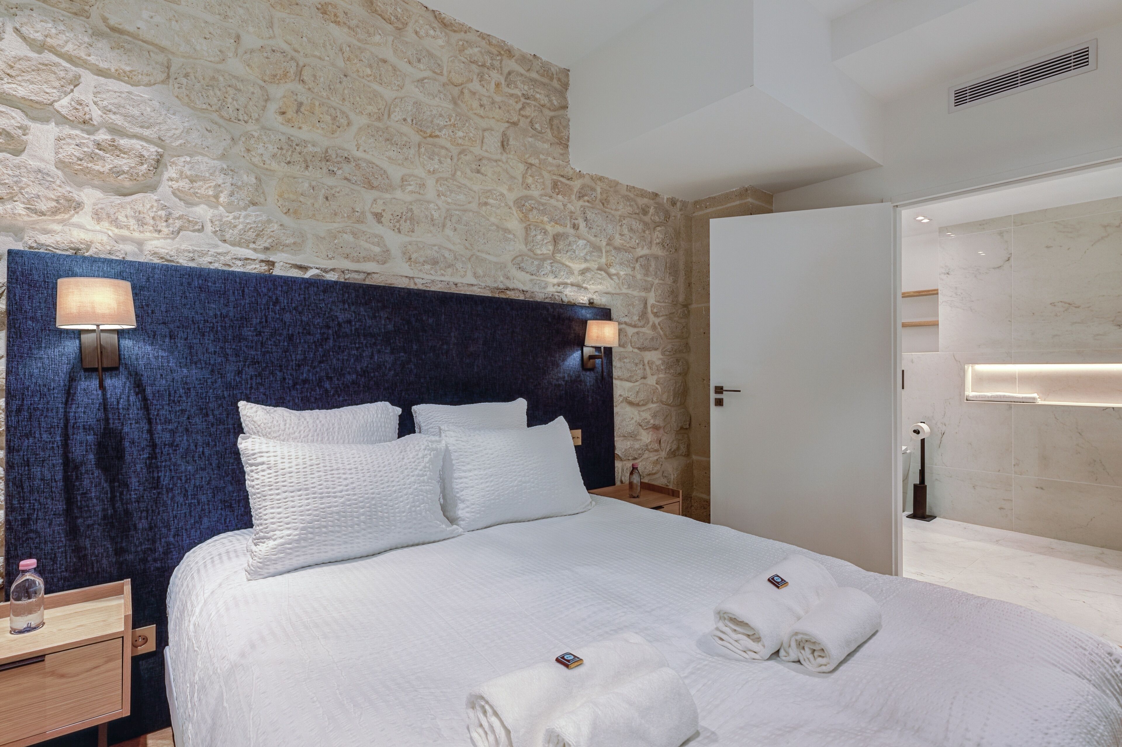 A neatly made bed with white linens and pillows, a stone wall behind, and a wooden nightstand with a lamp and a bottle.