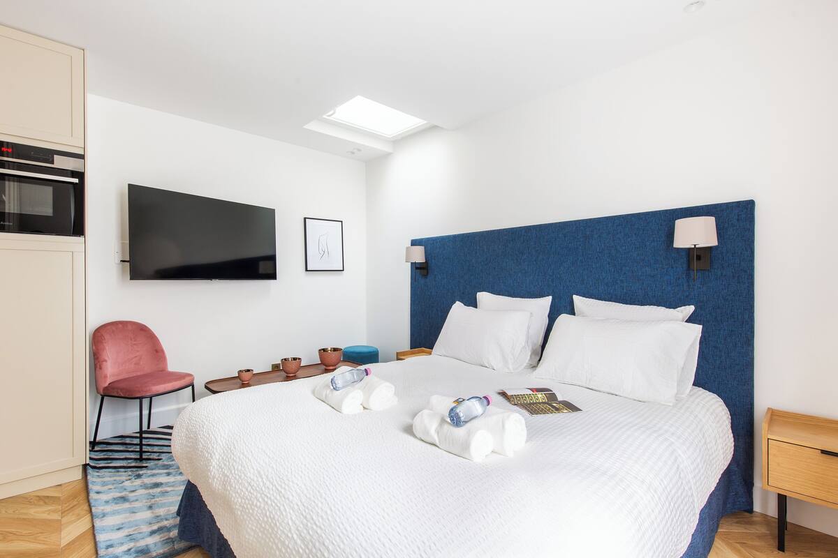 A modern hotel room with a large bed, a flat-screen TV, a blue headboard, and a small desk with a lamp.