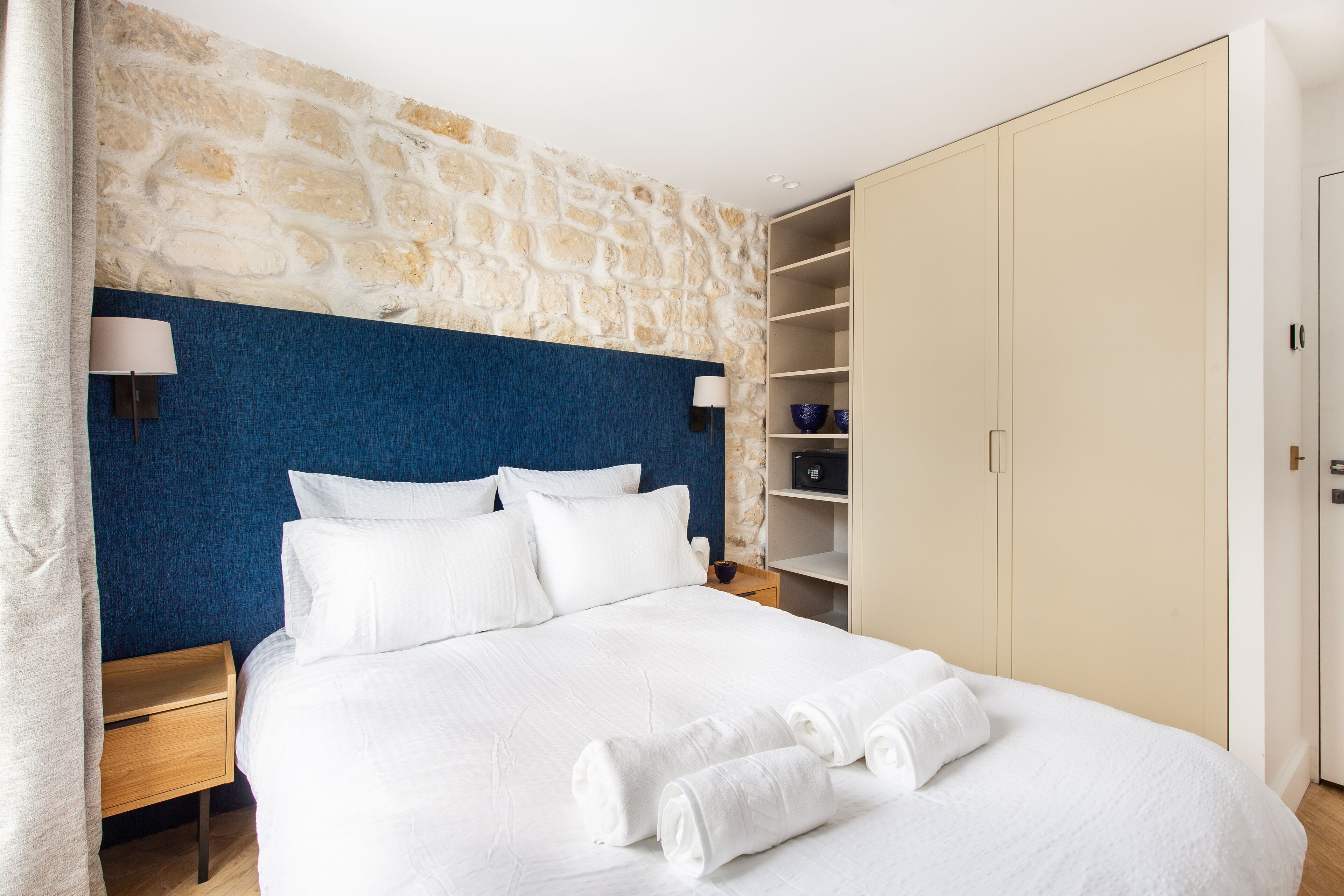 A neatly made bed with white linens and towels, a stone wall, a blue accent wall, and a wooden nightstand with a lamp.