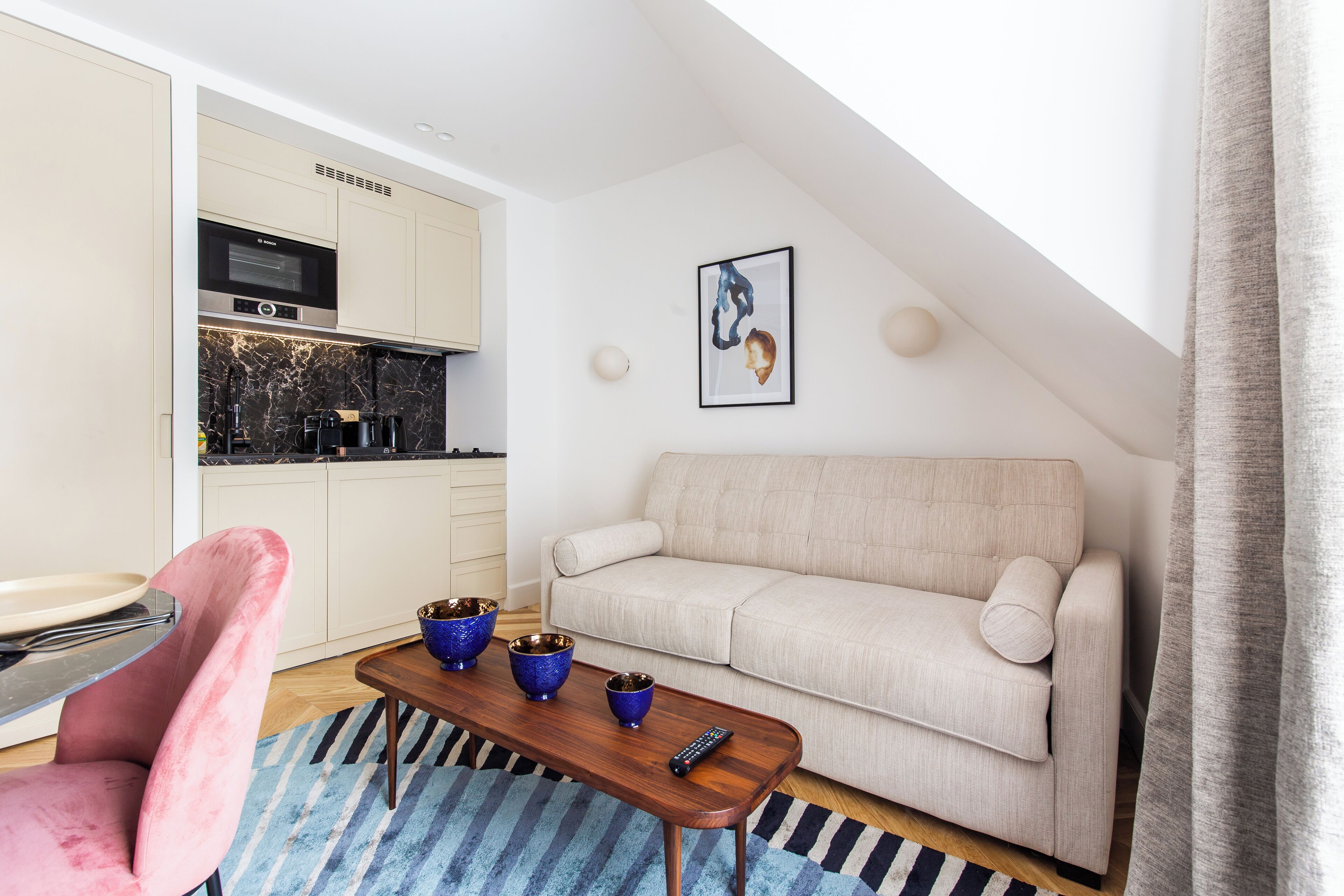 A modern living room with a sofa, a coffee table with blue bowls, a pink chair, and a kitchen in the background.