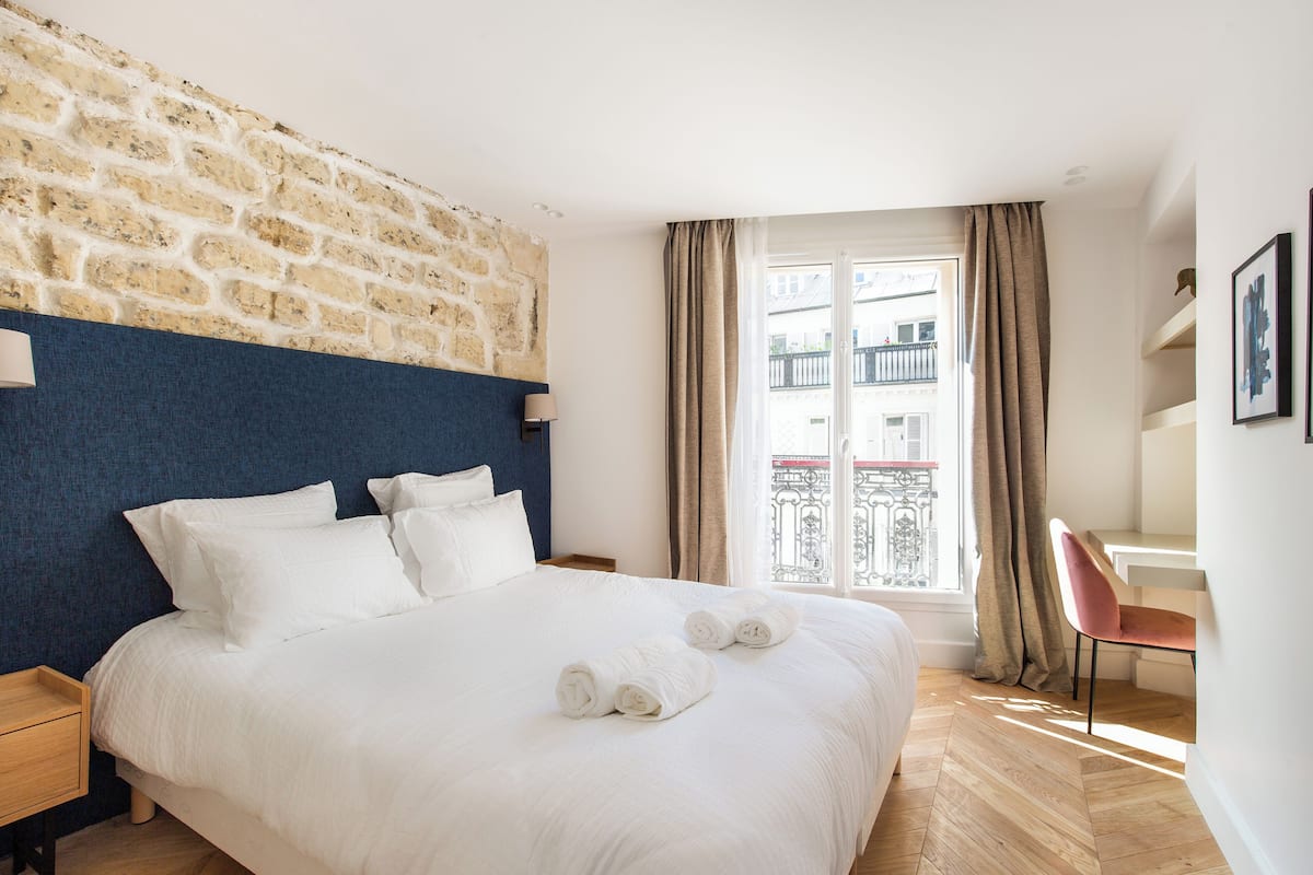 A hotel room with a brick wall, a bed with white linens, a desk with a chair, and a window with curtains.