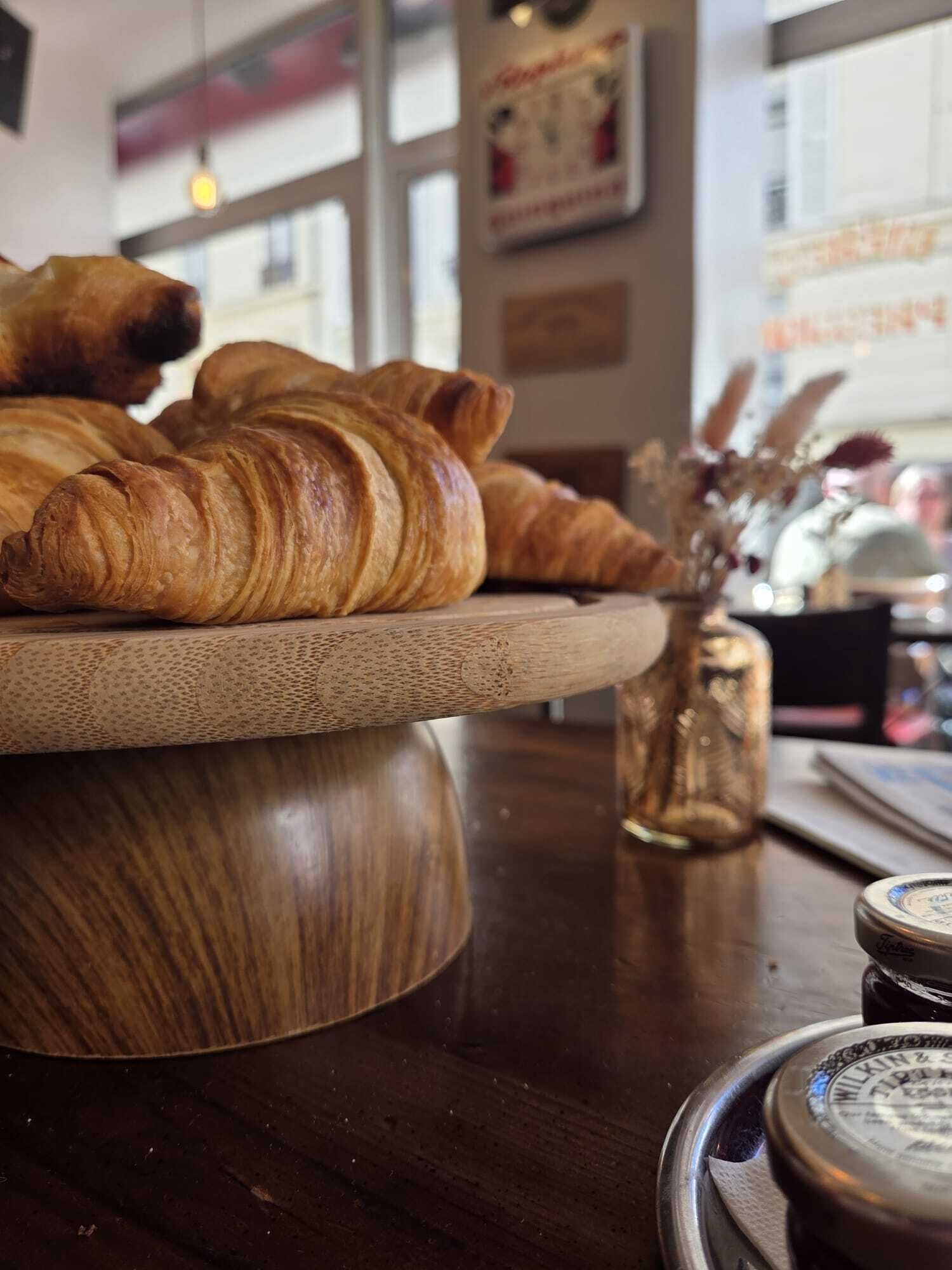 A wooden serving board with croissants and a glass jar on a table in a cafe setting.