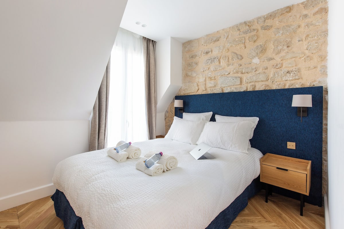 A neatly made bed with white linens and towels, a stone wall, and a wooden nightstand with a lamp.