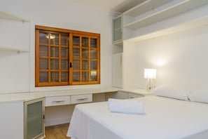 A bedroom with a bed, a desk, a chair, a window with blinds, and a nightstand with a lamp.