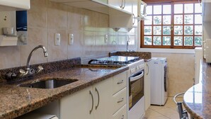A kitchen with white cabinets, a granite countertop, a built-in oven, a dishwasher, and a window with a view of trees. - LOCAR-IN GRAMADO- Leonardo da Vinci (Gramado)