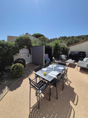 Un espace extérieur aménagé en terrasse, avec une table dressée pour les repas, entourée de chaises, et un barbecue à l’arrière-plan.