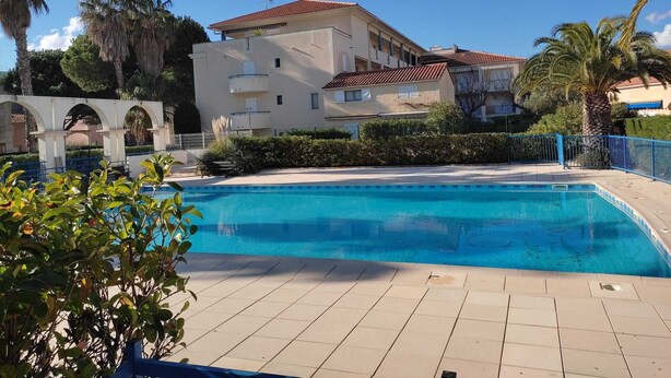 A swimming pool with a tiled deck, surrounded by greenery and a building with a red-tiled roof.