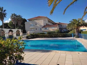 Piscina com deck revestido de azulejos, cercada por vegetação e um edifício com telhado de telhas vermelhas.