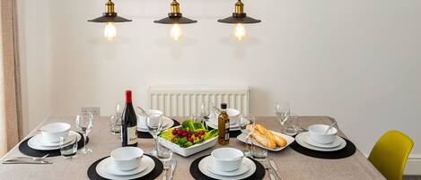 Mesa de jantar posta com pratos, copos, talheres e uma garrafa de vinho, com partituras emolduradas na parede acima.