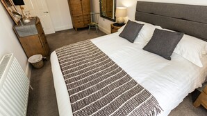 A bedroom with a bed, a dresser, a chair, a mirror, and a lamp. - Saint James By The Quay (Ilfracombe)