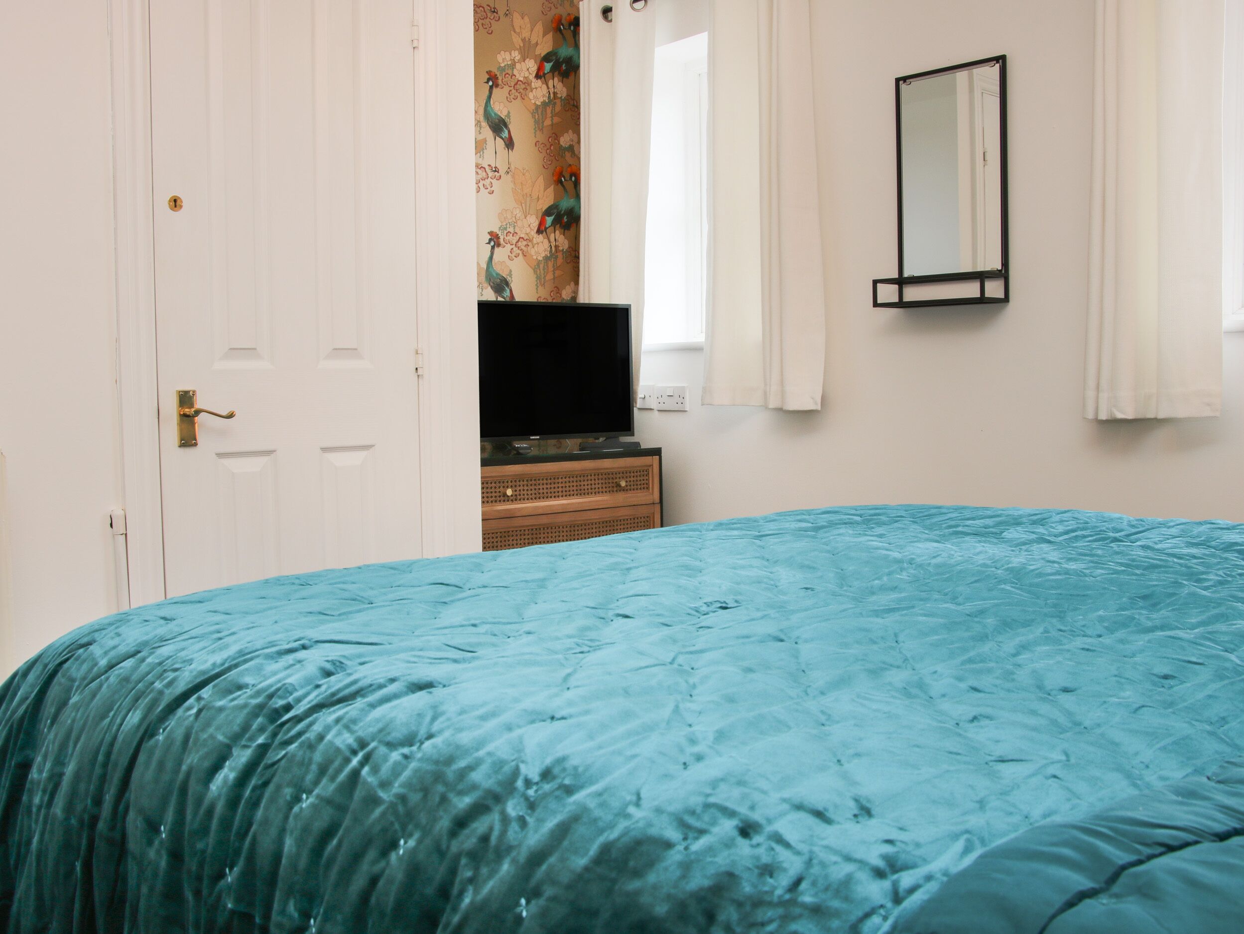 Quarto com colcha azul-turquesa, televisão sobre suporte de madeira, porta branca com puxador de latão e espelho na parede.