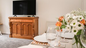 A living room with a flat-screen TV on a wooden cabinet, a dining table set with plates, glasses, and a vase with flowers. - Pippin's Place (Shrewsbury)